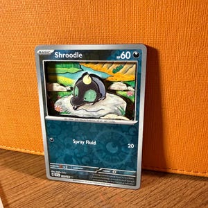 May include: A Pokémon trading card featuring a 3D Shroodle design. The card has a blue and silver border with the name "Shroodle" and "HP 60" printed on it. The card also features the text "Spray Fluid" and the number "20".