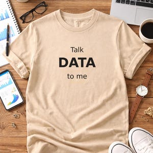 Puede incluir: Una camiseta beige con el texto "Talk DATA to me" en negro. La camiseta está sobre una superficie de madera con un portátil, una taza de café, un reloj, zapatillas y otros útiles de oficina. Es una prenda informal.