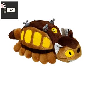 Puede incluir: Un peluche con forma de oruga marr&oacute;n y beige con detalles amarillos. El juguete tiene una cara sonriente y peque&ntilde;os detalles grises en la espalda. El texto "OTAKU DESK" est&aacute; en la esquina superior izquierda.