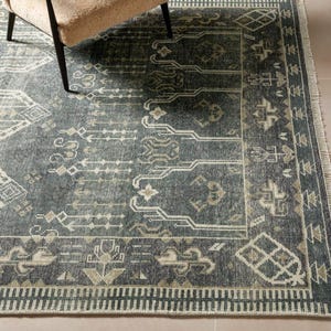 Peut inclure: Un tapis rectangulaire bleu canard avec un motif géométrique et une bordure crème. Le tapis présente un design vieilli avec diverses formes et lignes crème et beige. Une partie d'un fauteuil beige est visible en haut à gauche.