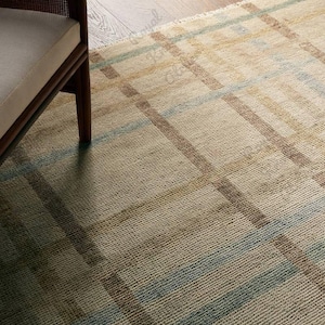 Op de afbeelding: Een geweven vloerkleed in beige met een rasterpatroon van bruine, blauwe en gele lijnen. Het kleed heeft een getextureerd oppervlak en is gedeeltelijk zichtbaar met een houten stoelpot. Het ontwerp bestaat uit kruisende lijnen.
