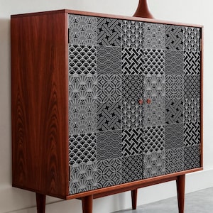 May include: A mid-century modern wooden cabinet with a geometric black and white patterned front. The cabinet has four tapered legs and two small, round copper-colored knobs. A brown lamp with a tan shade sits on top.