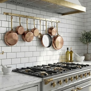Puede incluir: Una cocina con azulejos blancos de metro, una estufa de acero inoxidable y latón, y ollas de cobre colgadas. Botellas de aceite de oliva, un mortero y una maja, y plantas en macetas complementan la decoración. La estética es limpia y clásica.