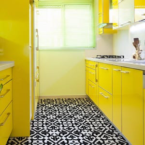 Puede incluir: Una cocina de color amarillo brillante con armarios y electrodomésticos a juego. El suelo presenta un diseño de baldosas con dibujos en blanco y negro. Se ve una ventana con persianas verdes.
