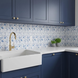 Puede incluir: Una cocina con gabinetes azul marino, una encimera blanca y un fregadero blanco de granja. El salpicadero presenta un patrón floral azul y blanco. Un grifo dorado y pequeñas plantas en macetas añaden detalles.