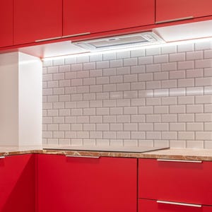 Könnte beinhalten: Moderne Küchengestaltung mit leuchtend roten Schränken und eleganten silbernen Griffen. Der Fliesenspiegel ist aus weißen U-Bahn-Fliesen, und eine Dunstabzugshaube aus Edelstahl ist über der Arbeitsplatte installiert. Ein LED-Streifen beleuchtet den Bereich.