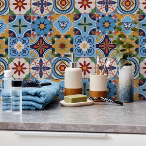 Puede incluir: Una escena de baño con un salpicadero de azulejos estampados en azul, amarillo y rojo. Sobre la encimera hay artículos de aseo, como una toalla azul, jabón, botellas, un cepillo y un portacepillos. También hay un jarrón con vegetación.