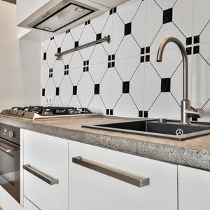 May include: Modern kitchen with white cabinets, a black sink, and a stainless steel faucet. The countertop is a speckled gray, and the backsplash features a black and white geometric tile pattern. A gas cooktop and oven are also visible.