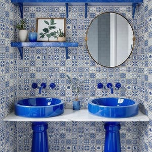 May include: A bathroom with blue and white patterned tile walls and floor. Two blue pedestal sinks sit on a white countertop, with a round mirror and shelves above. Decorative items include plants and vases.