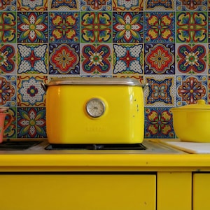 Op de afbeelding: Een gele broodrooster, pot en kopje staan op een gele aanrecht. De achtergrond bestaat uit kleurrijke tegels met patronen. De broodrooster heeft een temperatuurmeter. Een oranje kopje met een groen bloemmotief is ook zichtbaar.