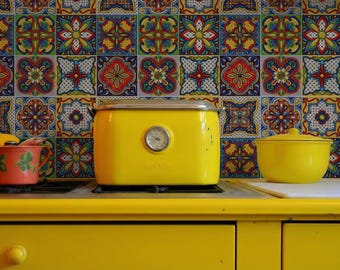 Puede incluir: Una tostadora, una olla y una taza amarillas están sobre una encimera amarilla. El fondo presenta azulejos estampados de colores. La tostadora tiene un indicador de temperatura. También se ve una taza naranja con un diseño floral verde.