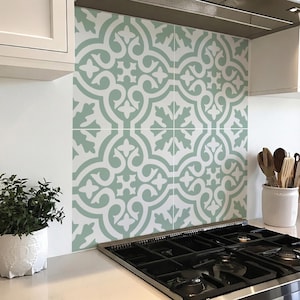 Może przedstawiać: Płytki kuchenne z powtarzającym się wzorem jasnozielonych i białych płytek dekoracyjnych. Płytki są zainstalowane nad czarną płytą gazową i obok białej szafki. Mała roślina doniczkowa stoi na blacie.