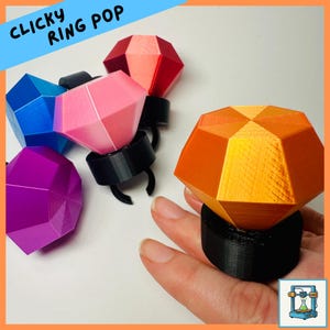 May include: A collection of colorful, faceted ring pops in various colors, including blue, pink, purple, red, and orange. The rings have a black base and are displayed on a white surface. The text "CLICKY RING POP" is in the top left corner.