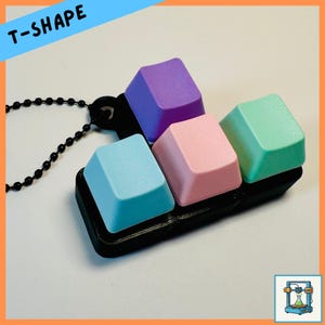 May include: A necklace pendant featuring four colorful keyboard keycaps in pastel shades of blue, pink, green, and purple. The keycaps are arranged on a black base and attached to a black ball chain. The word "T-SHAPE" is written in white letters on a blue background.