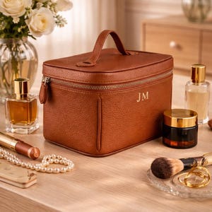 May include: A brown leather cosmetic bag with a top handle and a zipper closure. The initials "JM" are embossed on the front. The bag is surrounded by perfume bottles, makeup brushes, and jewelry on a wooden surface.
