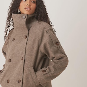 May include: A brown wool coat with a high collar and double-breasted button closure. The coat features two front pockets and button details on the sleeves. The coat is a neutral color and appears to be made of a soft material.