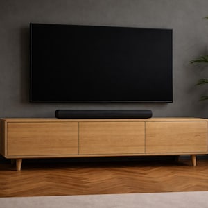 May include: A modern entertainment center featuring a large black television mounted on a gray wall. Below, a light wood cabinet with three doors holds a black soundbar. A potted plant adds a touch of greenery.