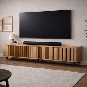 Puede incluir: Una moderna sala de estar con un televisor negro grande montado sobre un mueble de madera con detalles de listones verticales. Una barra de sonido negra se encuentra en el mueble. La habitación incluye un sofá, una alfombra y una planta en maceta.