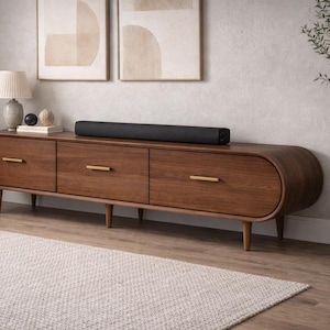May include: A walnut-stained media unit with three drawers and brass handles. A black soundbar rests on top. The unit is set against a neutral wall with framed artwork and a rug. The unit is approximately 180 cm long.