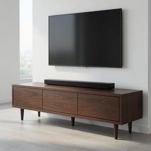 Può includere: Un mobile TV in legno marrone scuro con tre cassetti e gambe affusolate supporta una soundbar nera. Un grande televisore a schermo piatto nero è montato sulla parete bianca sopra. La stanza ha un design minimalista con una camera da letto visibile sullo sfondo.