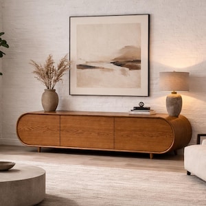 May include: A contemporary living room featuring a wooden media unit with three doors, a framed landscape artwork, and a round coffee table. A potted plant and a lamp are also visible. The room has a neutral colour scheme and a rug.