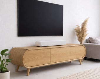 Könnte beinhalten: Ein modernes Wohnzimmer mit einem schwarzen Fernseher an einer weißen Wand. Darunter ein helles Unterhaltungsmöbel aus Eichenholz mit drei Türen und abgerundeten Kanten. Eine weiße Soundbar liegt auf dem Möbel. Ein beiger Teppich und eine Topfpflanze vervollständigen die Szene. Das Möbel ist ca. 180 cm lang.