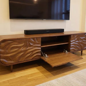 K&ouml;nnte beinhalten: Ein TV-Schrank aus Holz im Mid-Century-Modern-Stil mit geschwungenem Design und geschnitztem Wellenmuster auf den T&uuml;ren. Der Schrank hat ein offenes Regal und eine teilweise ge&ouml;ffnete T&uuml;r. Der Schrank ist braun, mit einer schwarzen Soundbar oben.