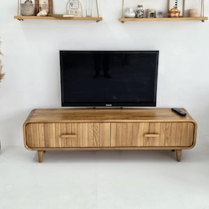 May include: A light brown wooden TV stand with two drawers and a ribbed front. The stand has rounded corners and sits on short, angled legs. A black flat-screen TV is mounted above the stand. A remote control rests on the stand's surface.