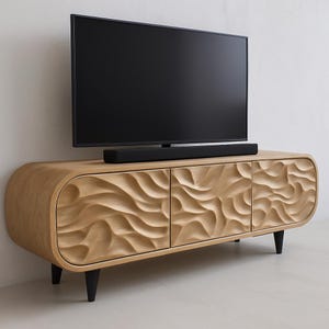 Könnte beinhalten: TV-Schrank aus hellem Holz mit drei Türen und einem geschnitzten Wellenmuster. Der Schrank hat schwarze Beine und eine abgerundete Form. Eine schwarze Soundbar befindet sich unter einem großen schwarzen Fernsehbildschirm.