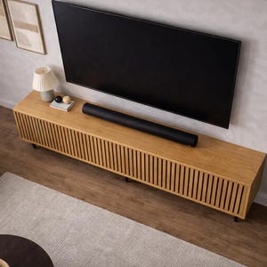 May include: A modern living room setup featuring a large black television mounted on a light gray wall. Below the TV is a light wood entertainment center with vertical slat detailing. A black soundbar rests on the center. A small lamp and plant add to the decor.