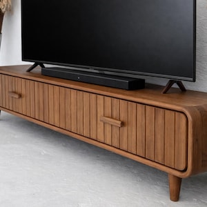 Può includere: Mobile TV in legno con finitura marrone scuro. Il mobile ha due cassetti con dettagli a scanalature verticali e bordi arrotondati. Una soundbar nera è posizionata sopra, sotto un grande televisore nero.