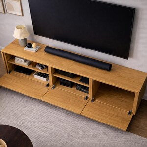 May include: A light-colored wooden TV stand with multiple compartments and doors, beneath a large black television. A black soundbar sits on top. A lamp, books, and decorative items are on the stand. A potted plant is to the right.