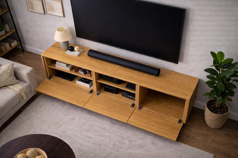 May include: A light-colored wooden TV stand with multiple compartments and doors, beneath a large black television. A black soundbar sits on top. A lamp, books, and decorative items are on the stand. A potted plant is to the right.