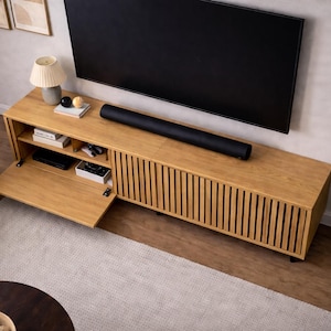 May include: A light wood entertainment center with a black soundbar and a large black television. The cabinet has a slatted front design and an open compartment. A potted plant and a lamp are also visible.