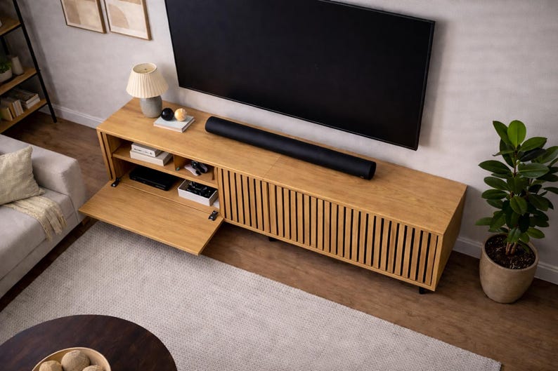 May include: A light wood entertainment center with a black soundbar and a large black television. The cabinet has a slatted front design and an open compartment. A potted plant and a lamp are also visible.