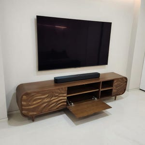 K&ouml;nnte beinhalten: Ein moderner TV-Schrank aus Holz mit einem wellenf&ouml;rmigen Design auf den T&uuml;ren. Der Schrank hat offene Regale und eine Soundbar. Ein gro&szlig;er, schwarzer Flachbildfernseher ist an der wei&szlig;en Wand montiert.
