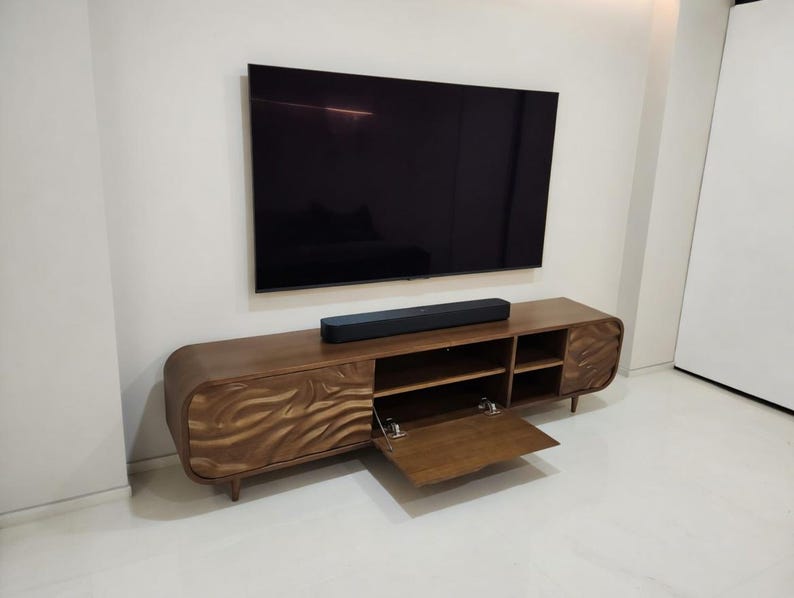 K&ouml;nnte beinhalten: Ein moderner TV-Schrank aus Holz mit einem wellenf&ouml;rmigen Design auf den T&uuml;ren. Der Schrank hat offene Regale und eine Soundbar. Ein gro&szlig;er, schwarzer Flachbildfernseher ist an der wei&szlig;en Wand montiert.