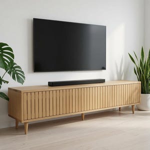 May include: A long, light-toned wooden media console featuring vertical slatted cabinet doors and tapered legs. A black soundbar sits on top of the console, positioned directly beneath a large, rectangular, black flat-screen television mounted on the wall.
