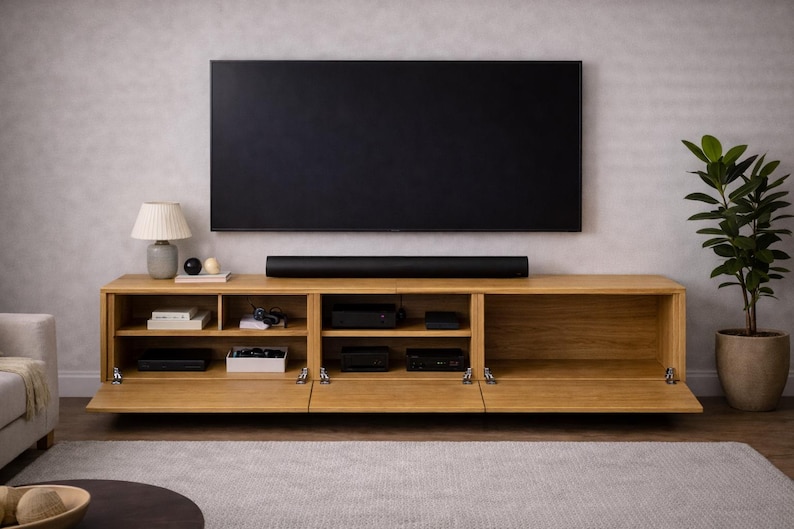 May include: A modern, light oak entertainment center with a large black television mounted above. The cabinet has open shelving and doors, with a soundbar below the TV. A lamp and plant add decor.