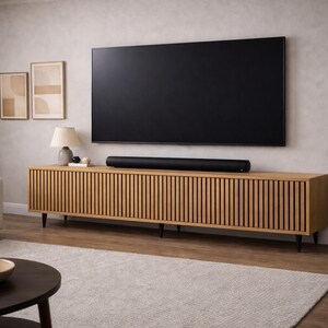 May include: A modern living room setup featuring a large black television mounted above a wooden cabinet with vertical slat detailing. A black soundbar sits on the cabinet. The room includes a sofa, rug, and a potted plant.
