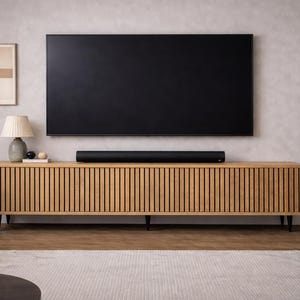 May include: A modern living room setup featuring a large black television mounted above a wooden cabinet with vertical slat detailing. A soundbar sits on the cabinet. A lamp, artwork, and a potted plant complete the decor.