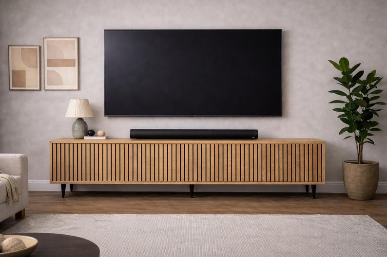 May include: A modern living room setup featuring a large black television mounted above a wooden cabinet with vertical slat detailing. A soundbar sits on the cabinet. A lamp, artwork, and a potted plant complete the decor.