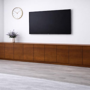 Puede incluir: Un mueble de televisi&oacute;n largo y bajo de madera con m&uacute;ltiples puertas de armario. Encima, un televisor de pantalla plana negro grande. Un reloj con borde dorado y un jarr&oacute;n de lavanda est&aacute;n en la pared encima del mueble. La habitaci&oacute;n tiene suelo gris claro y paredes blancas.