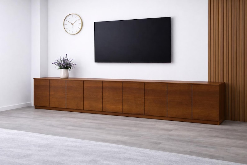 Puede incluir: Un mueble de televisi&oacute;n largo y bajo de madera con m&uacute;ltiples puertas de armario. Encima, un televisor de pantalla plana negro grande. Un reloj con borde dorado y un jarr&oacute;n de lavanda est&aacute;n en la pared encima del mueble. La habitaci&oacute;n tiene suelo gris claro y paredes blancas.