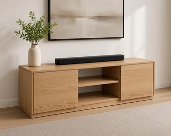 Puede incluir: Una consola multimedia de madera de roble claro con dos armarios laterales de puertas sólidas y una unidad central de estantería abierta con un estante. Una barra de sonido negra descansa sobre la superficie junto a un jarrón blanco.