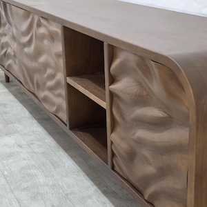 K&ouml;nnte beinhalten: Ein langes, niedriges Holzm&ouml;bel mit einzigartigem Design. Der Schrank hat eine hellbraune Oberfl&auml;che und weist ein wellenf&ouml;rmiges, strukturiertes Muster auf den T&uuml;ren auf. Er hat offene Regale und eine geschwungene Oberseite, ideal f&uuml;r ein Wohn- oder Unterhaltungsbereich.