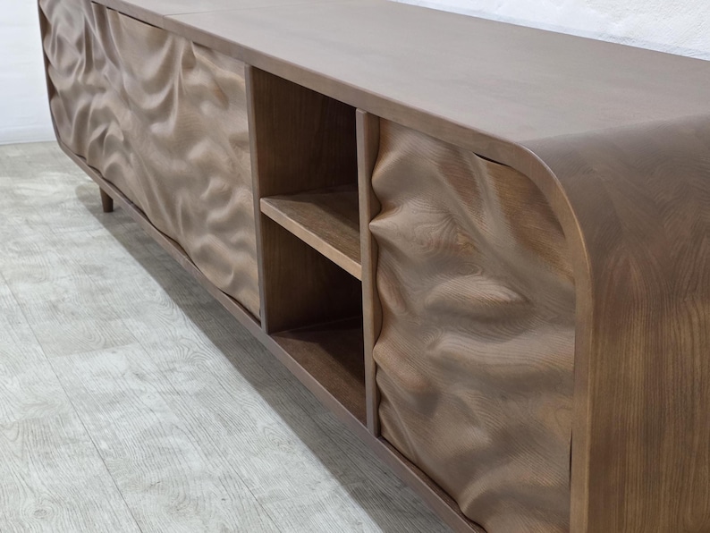 K&ouml;nnte beinhalten: Ein langes, niedriges Holzm&ouml;bel mit einzigartigem Design. Der Schrank hat eine hellbraune Oberfl&auml;che und weist ein wellenf&ouml;rmiges, strukturiertes Muster auf den T&uuml;ren auf. Er hat offene Regale und eine geschwungene Oberseite, ideal f&uuml;r ein Wohn- oder Unterhaltungsbereich.