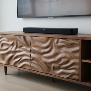 May include: A wooden media console with a carved wave design on the cabinet doors. A black soundbar sits on top of the console, below a mounted television. The console has a natural wood finish and a minimalist design.