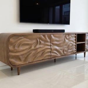 K&ouml;nnte beinhalten: Ein TV-Schrank aus Holz mit einem einzigartigen, geschnitzten Wellendesign auf den T&uuml;ren. Der Schrank hat eine warme, braune Oberfl&auml;che, abgerundete Kanten und ein kleines Regal. Er wird von vier konischen Beinen getragen. Eine schwarze Soundbar liegt oben.