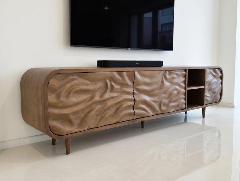 K&ouml;nnte beinhalten: Ein TV-Schrank aus Holz mit einem einzigartigen, geschnitzten Wellendesign auf den T&uuml;ren. Der Schrank hat eine warme, braune Oberfl&auml;che, abgerundete Kanten und ein kleines Regal. Er wird von vier konischen Beinen getragen. Eine schwarze Soundbar liegt oben.